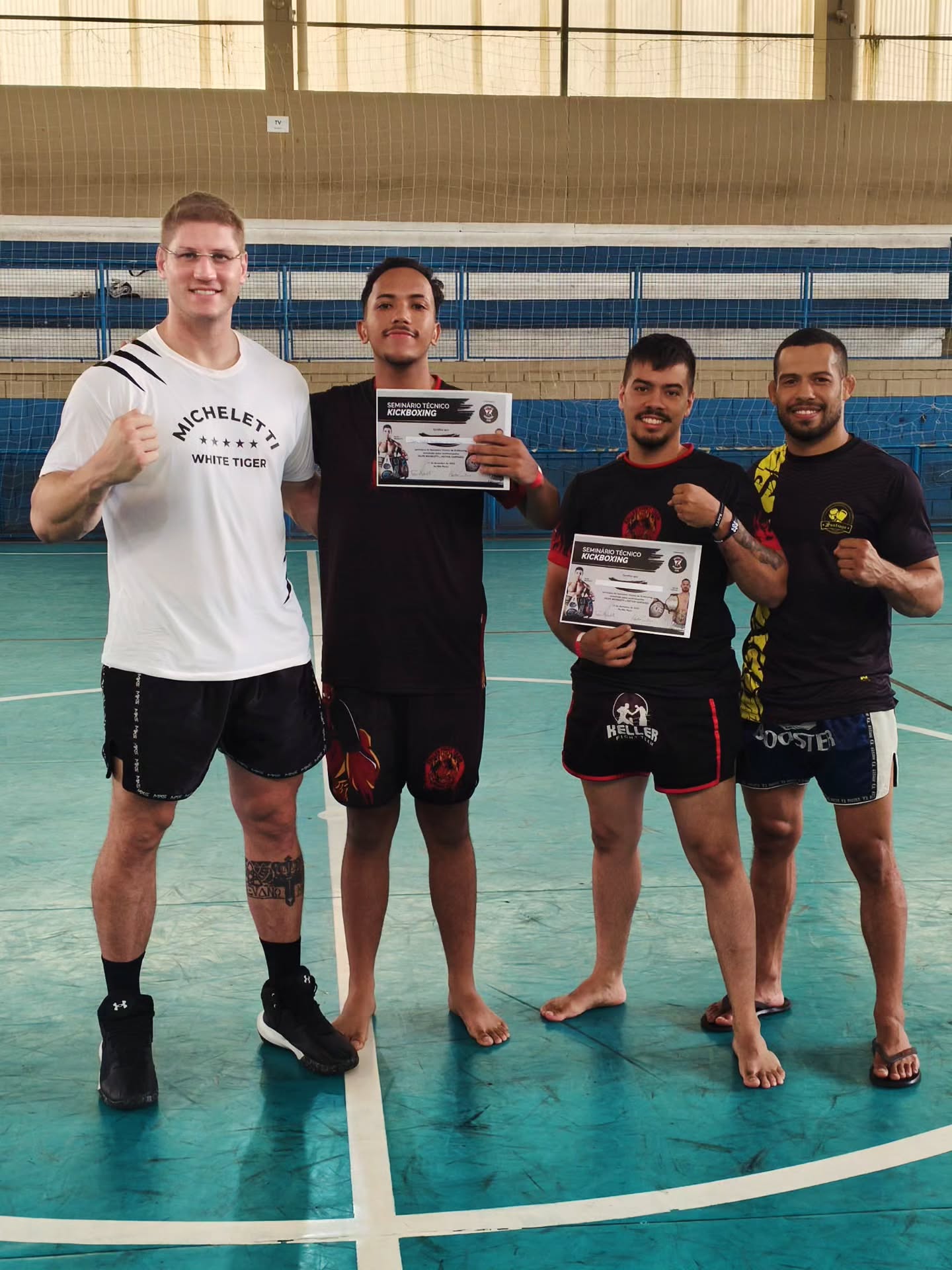 Quatro lutadores posam com certificados do seminário técnico de kickboxing em uma quadra poliesportiva.