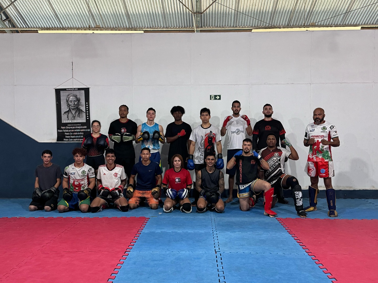 Grupo de alunos com luvas e proteções posa em um ginásio com tatame azul e vermelho, após treino especial.
